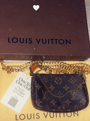 Louis Vuitton mini pochette bag.  - Säljer en liten ikonisk mini pochette väska från Louis Vuitton med klassiskt monogrammönster i brunt och beige. Väskan har guldfärgad kedja och dragkedja upptill.  Den har även ett medföljande crossbody axelband i guld så det går även att hänga den över axeln.  Det ingår även LV låda och LV dustbag.  The Mini Pochette Accessoires in Monogram canvas is a stylish way to carry essentials. Its secure zip protects a spacious compartment, while the chain and hook enable it to be attached to a larger b