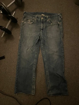 True Religion Bobby Super T jeans blå - Säljer ett par blå True Religion Bobby Super T jeans med grova vita sömmar och slitna detaljer. Klassiska bakfickor med broderad logga och råa kanter vid benslut. Jeansen har en loose passform och är tillverkade i jeansmaterial med coola metalldetaljer.