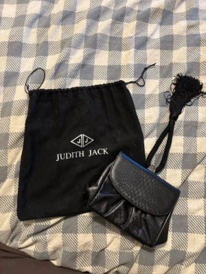 Svart kuvertväska Judith Jack - Snygg svart kuvertväska från Judith Jack med reptilmönstrad yta och dekorativ tofs i snöre. Väskan har magnetlås och insida med logotyp-tryck. Perfekt storlek för det viktigaste och en cool detalj till din outfit.