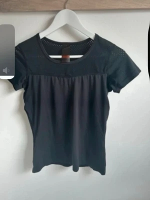 Svart kortärmad topp med meshdetaljer - Säljer en svart topp med korta ärmar och rund hals. Överdelen och ärmarna har meshpaneler med små hål som ger en sportig vibe. Materialet känns lätt och stretchigt, perfekt för dig som gillar stilrena och enkla plagg med en twist.