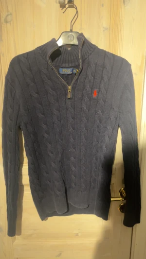 Mörkblå stickad tröja Polo Ralph Lauren - Säljer en mörkblå stickad tröja från Polo Ralph Lauren i storlek XS. Tröjan har kabelstickat mönster, halv dragkedja och en röd broderad logga på bröstet. Hög krage och långa ärmar ger en klassisk och snygg look.