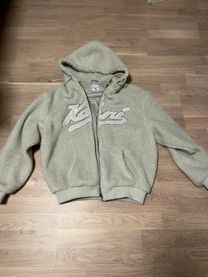 Beige teddyjacka från Karl Kani XS - Mjuk och fluffig teddyjacka i beige från Karl Kani, med huva och dragkedja framtill. Jackan har broderad logga på bröstet, ribbade muddar och två fickor. Perfekt för dig som gillar chill och cozy vibes.