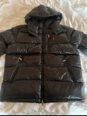 Svart pufferjacka från Polo Ralph Lauren - Svart pufferjacka med huva från Polo Ralph Lauren. Jackan har en röd logga på bröstet, flera dragkedjefickor och quiltad design. Materialet är glansigt och syntetiskt, perfekt för kalla dagar. Snygg och praktisk med en modern look.