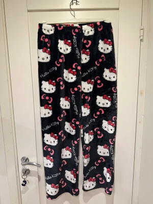 Hello Kitty mjukisbyxor svart/rosa - Supermysiga svarta mjukisbyxor med Hello Kitty-motiv och rosa detaljer. Byxorna har ett rakt snitt och är gjorda i mjuk fleece, perfekta för chill dagar hemma. Elastisk midja för extra komfort och ett gulligt all-over print med Hello Kitty och rosetter.