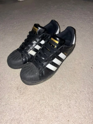 Adidas Superstar svarta sneakers - Svarta Adidas Superstar sneakers med klassiska vita ränder på sidorna och guldiga detaljer på plösen. Skorna har snörning, platt sula och rund tå. Ovandelen är i skinn och insidan är fodrad med textil. Perfekt för dig som gillar retrostil och streetwear.