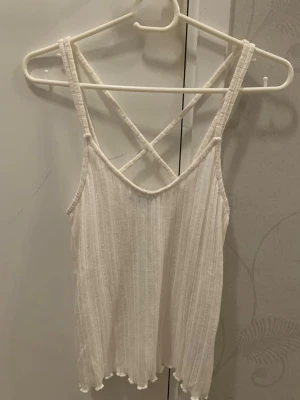 Vit ribbad linnetopp XXS - Säljer en vit ribbad linnetopp från Sinsay i storlek XXS men passar även XS. Toppen har smala axelband som korsas i ryggen och en lätt vågig kant nedtill. Är creme vit i färgen. Materialet är mjukt och stretchigt, perfekt för varma dagar. Enkel och fräsch design som är lätt att matcha med jeans eller shorts.