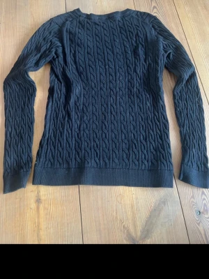 Svart kabelstickad tröja Basic You - Svart kabelstickad tröja från Basic You i ekologisk bomull. Tröjan har långärmad design och ribbade muddar vid ärmslut och nederkant. Snygg struktur med flätmönster över hela plagget, perfekt för dig som gillar klassisk stil.