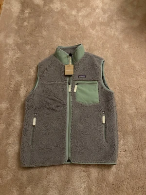 Grå fleeceväst från Patagonia - Snygg grå fleeceväst från Patagonia med ljusgrön krage och detaljer. Västen har tre fickor med dragkedja, varav en bröstficka i kontrasterande tyg. Perfekt för lager-på-lager och outdoor-häng.