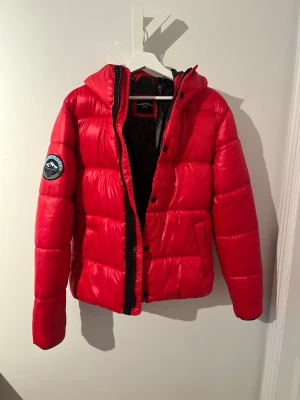 Röd pufferjacka från Superdry S - Snygg röd pufferjacka från Superdry med huva och svart innerfoder. Jackan har ett Superdry-märke på vänster ärm, praktiska fickor och stängs med både dragkedja och knappar. Perfekt för kalla dagar och har en cool, sportig vibe. Väldigt lite använd och i mycket bra skick!