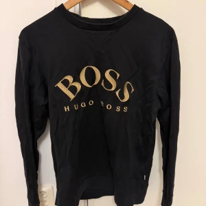 Svart långärmad tröja Hugo Boss - Svart långärmad tröja från Hugo Boss med stort guldigt BOSS-tryck på bröstet. Rund halsringning med ribbad kant och mjukt bomullsmaterial. Storlek M men känns som S.