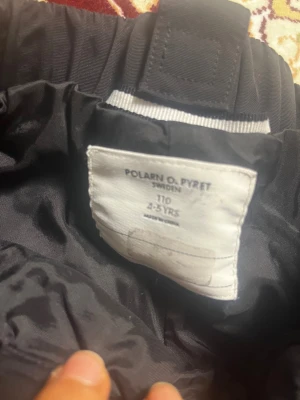 Svarta täckbyxor Polarn O. Pyret 110 - Svarta täckbyxor från Polarn O. Pyret i storlek 110. Byxorna har resår i midjan och benslut, samt reflexdetaljer på benen för extra synlighet. Perfekta för kalla dagar och utomhuslek. Klassisk design med slitstarka förstärkningar på knän och bak.