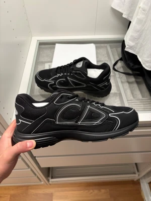 Svarta Dior B30 sneakers - Säljer ett par svarta Dior B30 sneakers med coola silvriga detaljer och CD-logga på sidan och hälen. Skorna har mesh och syntetmaterial, snörning och en chunky platt sula. Perfekt för dig som gillar exklusiv streetstyle och vill sticka ut med designer på fötterna.