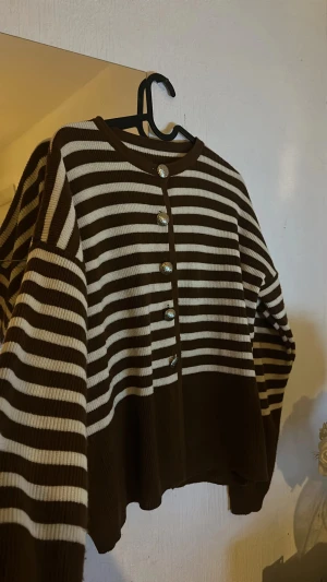 Randig brun kofta med knappar - Superfin brun och vit randig kofta med snygga silvriga knappar framtill. Koftan är stickad och har en loose passform med långa ärmar. Jag hittar ingen storlekslapp, men skulle uppskatta den till S/M.