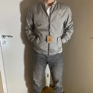 Burberry Windbreaker - Snygg ljusgrå windbreaker från Burberry med dragkedja framtill och ribbade muddar. Jackan har två fickor med knapp och diskret broderad logga på bröstet. Perfekt för dig som vill ha en stilren och klassisk look nu inför hösten Finns olika storlekar och tveka inte vid frågor 😊🍁