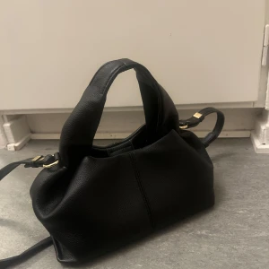 Svart axelväska i skinn med gulddetaljer - Snygg svart axelväska i mjukt skinn med stilrena gulddetaljer och breda handtag. Väskan har en enkel men modern design och en praktisk axelrem som gör den lätt att bära. Perfekt för dig som gillar en clean och tidlös look.