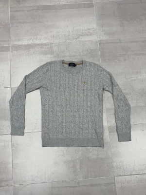 Grå kabelstickad tröja Morris Lady - Snygg grå kabelstickad tröja från Morris Lady i 100% pima bomull. Tröjan har rund hals, ribbade muddar och ett litet broderat emblem på bröstet. Perfekt för dig som gillar klassisk och stilren look med lite preppy vibe.