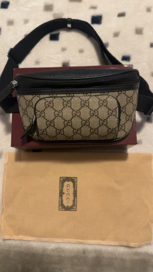 Gucci beige midjeväska med GG-mönster - Säljer en beige midjeväska från Gucci med klassiskt GG-mönster och svarta detaljer i skinn. Väskan har justerbart band, flera fack med dragkedja och levereras med original dustbag och box. Perfekt för dig som vill ha en ikonisk accessoar med street vibe.