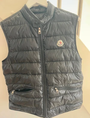  dunväst  Moncler - Svart dunväst från Moncler med quiltad design och hög krage. Västen har två fickor med dragkedja, Moncler-logga på bröstet och stängs med dragkedja framtill. Materialet är glansigt och västen har en clean, stilren look som funkar till många outfits. kvitto finns och påse inköpt på Moncler butiken i Stockholm. Nypris är över 9000kr. 