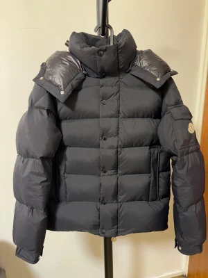 Svart Moncler pufferjacka med huva - Säljer en svart pufferjacka från Moncler med avtagbar huva och klassisk logga på ärmen. Jackan har både dragkedja och knappar framtill, samt praktiska fickor. Den är rejält vadderad och har en modern, boxig siluett som ger en cool look.