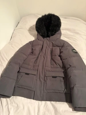 Grå pufferjacka Supply & Demand - Snygg grå pufferjacka från Supply & Demand med svart fuskpäls runt huvan. Jackan har två stora fickor med lock, två bröstfickor med dragkedja och en cool patch på ärmen. Perfekt för kalla dagar med sin tjocka vaddering och mjuka insida.