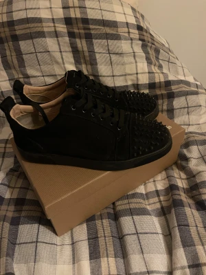 Christian Louboutin svarta sneakers med nitar - Svarta sneakers från Christian Louboutin med ikoniska röda sulor och coola nitar på tån. Skorna har snörning och är tillverkade i mocka och skinn, vilket ger en lyxig känsla. Perfekt för dig som vill sticka ut med en edgy och exklusiv stil. Båda skorna har tappat in spik var