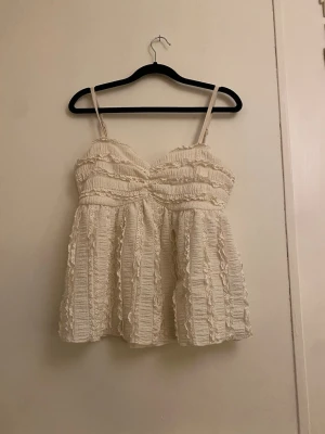 Krämvit volangig linnetopp med spets - Säljer en krämvit linnetopp med tunna axelband och volangdetaljer. Toppen har v-ringad skärning och är dekorerad med spets och rynkade partier som ger en romantisk och bohemisk vibe. Perfekt för varma dagar och sommarhäng.