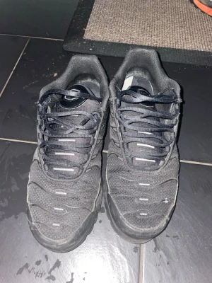 Svarta Nike Air Max Plus skor - Svarta Nike sneakers med mesh- och syntetmaterial, coola vågformade detaljer på ovandelen och robust sula med Vibram-mönster. Skorna har snörning och en sportig vibe, perfekta för dig som gillar streetstyle och komfort.