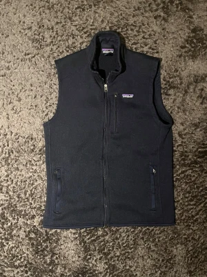 Mörkblå fleeceväst från Patagonia - Säljer en mörkblå fleeceväst från Patagonia i storlek M. Västen har hög krage, hel dragkedja framtill och tre praktiska fickor med dragkedja. Perfekt för lager-på-lager och chill stil. Diskret logga på bröstet för extra vibe.