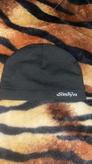 Svart och vit satinmössa från Satyn - Säljer 2 stilrena mössor från Satyn i svart och vit. Utsidan är slät och enkel med Satyn-logga broderad framtill. Insidan är fodrad med glansig satin som känns lyxig mot huden. Mössorna har en klassisk, rundad form och passar perfekt för dig som gillar minimalistisk stil.