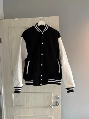 Exklusiv collagejacka med äkta läder - Säljer en klassisk baseballjacka i svart med vita ärmar och vita detaljer av äkta läder. Jackan har ribbade muddar och krage med marinblå och vita ränder, knäppning med vita knappar och två fickor fram. Fodret är quiltat och insidan har en snygg etikett. Perfekt för en sportig och cool look. Pris kan diskuteras, hör av dig vid minsta fråga!