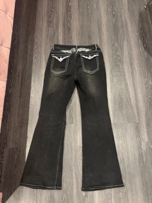 Mexikanska jeans - Svarta jeans med bootcut passform och markerade vita sömmar. Baksidan har broderade vingar och fickor med diamanter och silvriga knappar. Jeansen har mellan midja och är tillverkade i klassiskt denimtyg. Perfekta för dig som gillar statement-detaljer.