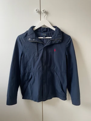 Vindjacka från Polo Ralph Lauren - Snygg marinblå vindjacka från Polo Ralph Lauren med röd broderad logga👌Jackan kostar 3200kr ny och mitt pris är 899kr!         Jackan har två hål på vardera sida vid framfickorna men det går att sy ihop🧵 Passar dig som är ca 168-175cm! Bara att fråga om du har andra funderingar👍👑