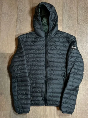 Svart pufferjacka från Colmar - Snygg svart pufferjacka från Colmar med huva och horisontella sömmar. Jackan har dragkedja framtill, två fickor med dragkedja och en diskret logga på ärmen. Perfekt för kyliga dagar och enkel att matcha med allt.