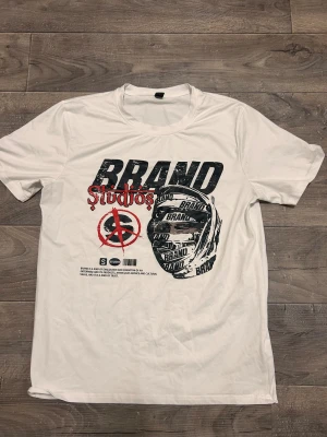 Vit t-shirt med tryck från Brand Studios - Vit t-shirt med coolt grafiskt tryck i svart och rött framtill. Trycket har texten 'Brand Studios' och ett ansikte insvept i band med ordet 'Brand'. T-shirten har rund hals och är kortärmad, perfekt för en streetwear-look.
