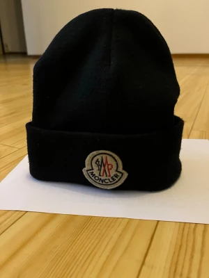 Svart Moncler mössa med logga - Svart mössa från Moncler med klassisk uppvikt kant och broderad Moncler-logga framtill. Mössan är stickad i mjukt material och har en liten vit etikett med Moncler-tryck på sidan. Perfekt för kalla dagar och snygg till streetwear.