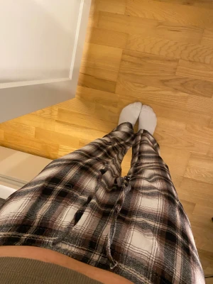 Rutig mjukisbyxa med snörning - Säljer ett par rutiga mjukisbyxor i svart, vitt och brunt. Byxorna har en avslappnad passform och dragsko i midjan för extra komfort. Perfekta för chill dagar hemma eller när du vill vara bekväm men ändå se snygg ut.