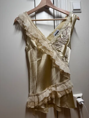 Champagnefärgad blus från Reclaimed Vintage - Supersöt blus i champagnefärgad satin från Reclaimed Vintage. Den har v-ringning, broderade detaljer med pärlor och volanger i spets längs kanterna. Blusen är ärmlös och har en omlott-look som ger en romantisk vibe.