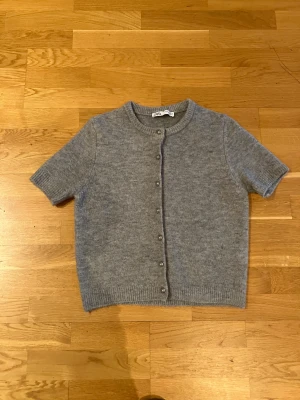 Grå kortärmad kofta från Zara - Säljer en grå, mjuk och stickad kofta från Zara med korta ärmar och rund hals. Koftan har knappar framtill och ribbade kanter vid ärmslut och nederkant. Perfekt för lager-på-lager och chill stil.