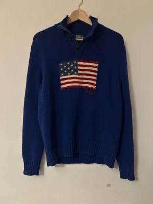 Blå stickad tröja Polo Ralph Lauren - Snygg blå stickad tröja från Polo Ralph Lauren med amerikansk flagga på bröstet och RL67-broderi. Tröjan har halv dragkedja, ribbade muddar och lång ärm. Perfekt för dig som vill sticka ut med en klassisk och cool look. Tröjan är i storlek XL men är liten i storleken så det passar S/M.
