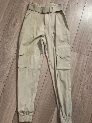 Beige cargopants med bälte - Snygga beige cargopants med elastiska benslut och praktiska fickor på sidorna. Byxorna har ett matchande bälte i midjan och är tillverkade i ett lätt syntetmaterial som ger en cool och avslappnad look. Använda enstaka gång, som nya! Nypris 800 