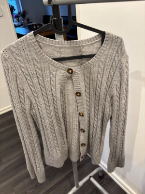 Grå kabelstickad kofta med knappar - Snygg grå kabelstickad kofta med guldfärgade knappar framtill. Koftan har rund halsringning, långa ärmar och ribbade muddar vid ärmslut och nederkant. Perfekt för dig som gillar klassisk stil med en twist.
