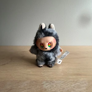 Secret Labubu - Supersöt liten gosedjur, formad som en fluffig grå figur med regnbågsfärgade ögon och öron. Kort och låda medföljer. Pälsig yta och svart ring baktill för att fästa på väska eller nycklar. Perfekt accessoar för dig som gillar gulliga detaljer.