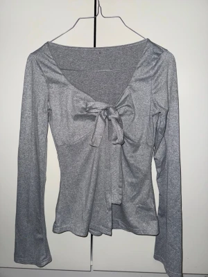 Grå omlottopp med öppning och knyt framtill - Snygg grå omlottopp med lång ärm och v-ringning. Toppen har en öppning och ett knyt framtill som ger en cool detalj och sitter figurnära. Materialet är mjukt och stretchigt, perfekt för att skapa en chill men ändå stilren look.