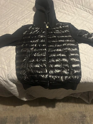 Svart cardigan  Moncler - Svart cardigan från Moncler med glansig quiltad front och ärmar i mjuk bomull. Jackan har huva med snörning, dragkedja framtill och en snygg Moncler-logga på ärmen. Praktiska fickor med dragkedja och coolt streetwear-vibe.