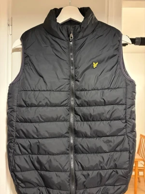 Svart dunväst från Lyle & Scott - Svart dunväst från Lyle & Scott med gul broderad logga på bröstet. Västen har quiltad design, hög krage och dragkedja framtill. Perfekt att slänga över en hoodie för extra värme. Snygg och enkel att matcha med olika stilar.
