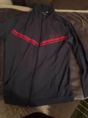 Blå ziptröja från Hugo Boss XL - Snygg marinblå ziptröja från Hugo Boss med röda ränder över bröstet och logga. Tröjan har lång ärm, hög krage och hel dragkedja framtill. Perfekt för dig som gillar sportig och stilren look. Materialet är mjukt och lätt polyester.