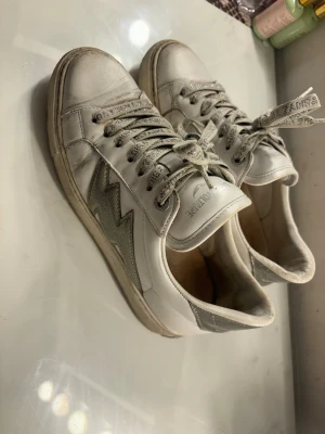 Zadig & Voltaire vita sneakers med blixt - Snygga vita sneakers från Zadig & Voltaire med silvriga detaljer och glittrande snören med logotyp. Skorna har en cool blixt på sidan i silver och platt sula. Tillverkade i skinn med textil på snörningen och glitter på hälen. Perfekt för dig som gillar statement-skor. Nypris 1900kr