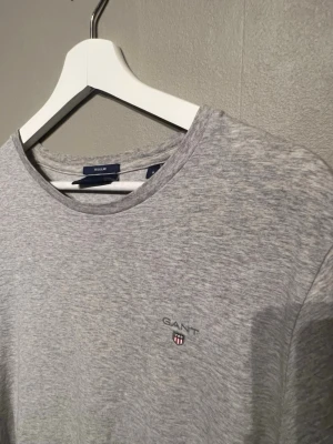 Grå t-shirt från GANT med logga - Snygg gråmelerad t-shirt från GANT med rund hals och diskret logga broderad på bröstet. Klassisk passform och mjukt bomullsmaterial som känns skönt mot huden. Perfekt för dig som gillar stilrena och enkla plagg med en touch av premium.