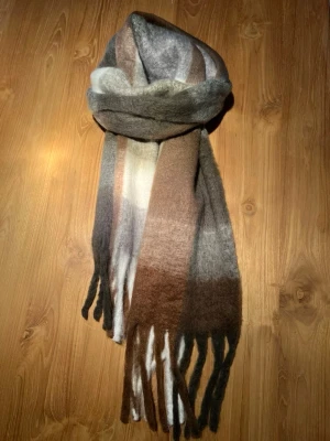 Rutig brun och grå halsduk med fransar - Mysig halsduk med rutigt mönster i bruna, gråa och vita toner. Den har långa fransar i ändarna och är tillverkad i ett mjukt, ulligt material som känns skönt mot huden. Perfekt att svepa runt halsen för extra värme och stil.