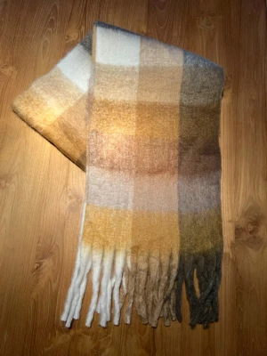 Rutig brun ullhalsduk - Säljer en mysig rutig halsduk som liknar acne  i mjuk ull med breda blockränder i beige, brunt, vitt och grått. Halsduken har långa fransar längs nederkanten som ger en chill vibe. Perfekt att slänga över en hoodie eller tröja när det är kallt ute.